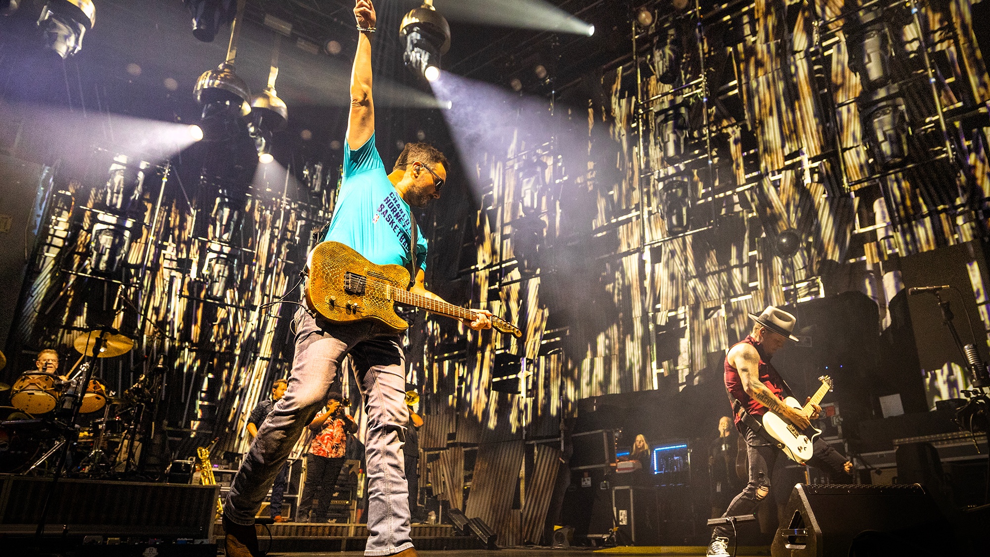 Country artist Eric Church holding hand up whilst playing electric guitar with band in the background against an interactive backdrop with handing lights