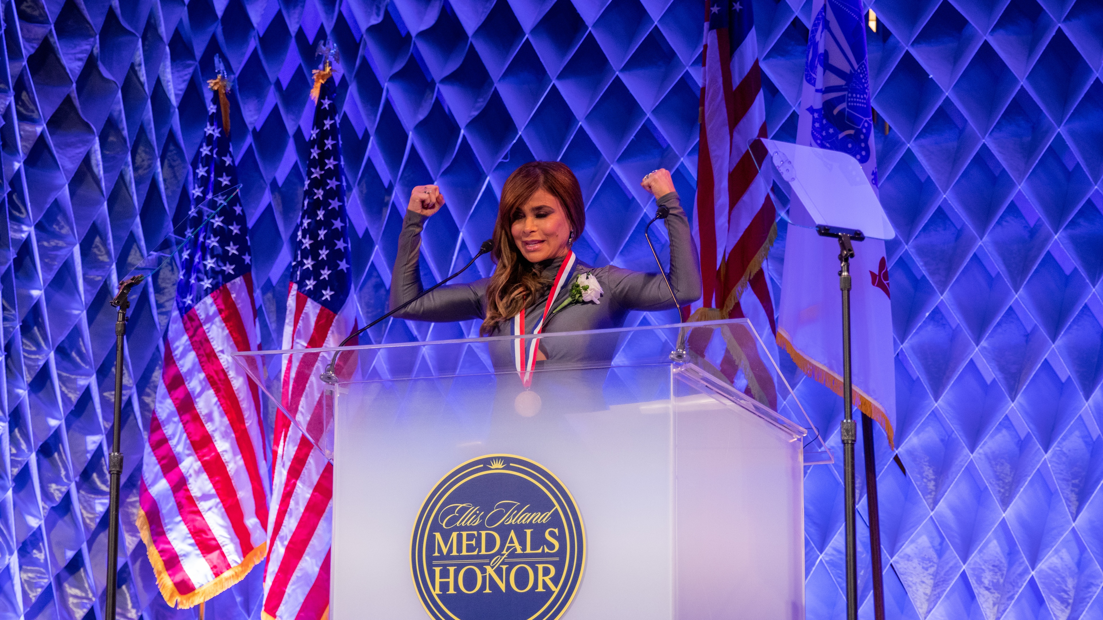 Woman wearing medal with arms raised stood at podium with geometric blue backdrop and american flags