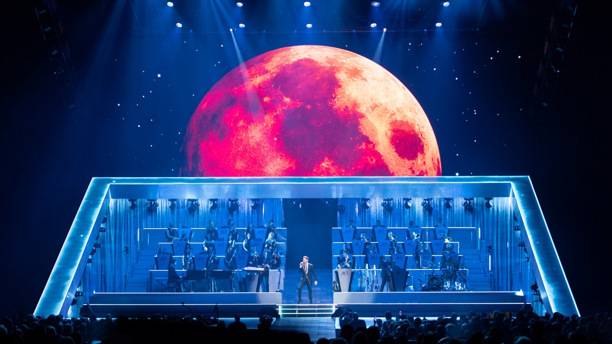 Michael Bublé performing on stage at The Higher Tour, seated orchestra to the sides, red moon in the sky backdrop