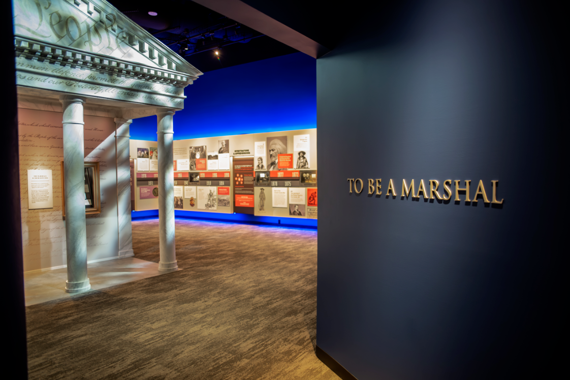 Museum room with To Be A Marshall in gold 3D lettering on a dark blue wall in the foreground, a replica marble building facade, and a historic timeline in the background