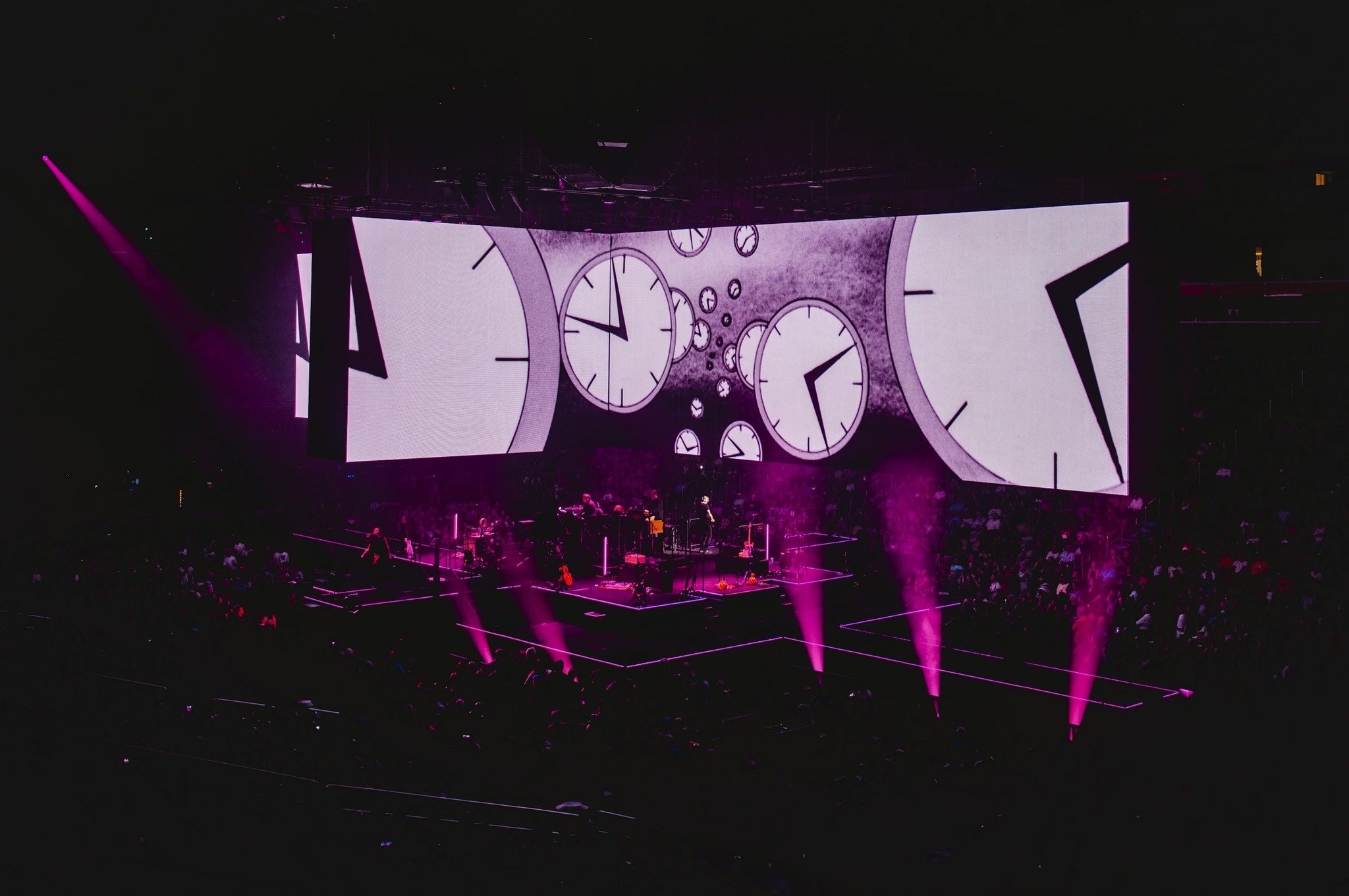 bird's eye view of Roger Waters performing during the This Is Not A Drill Tour, pink lights, screen above performers feature illustrations of analog clocks