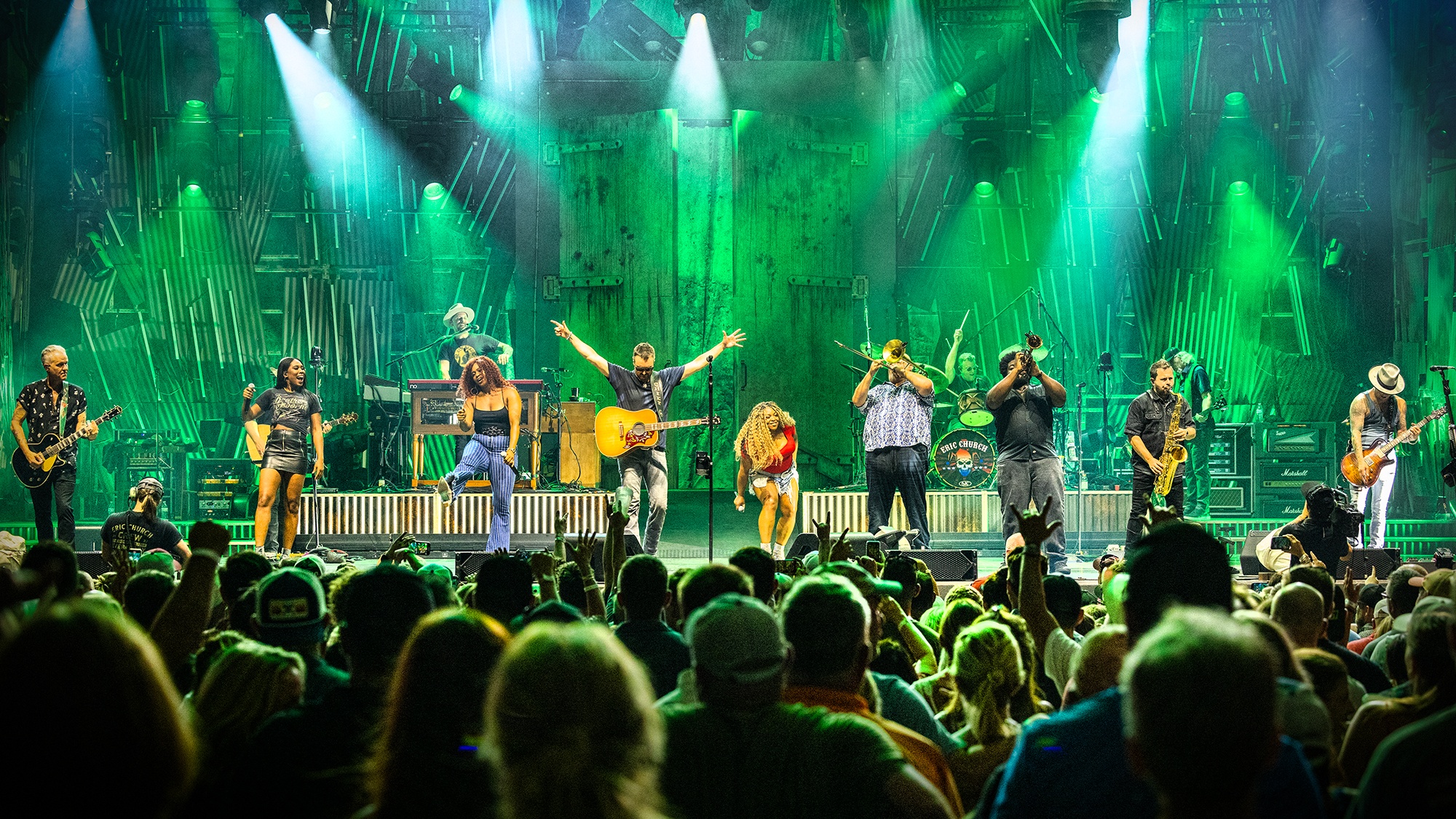 Country artist Eric Church and band stood in a line playing with crowd in front of green backdrop and with hanging lights