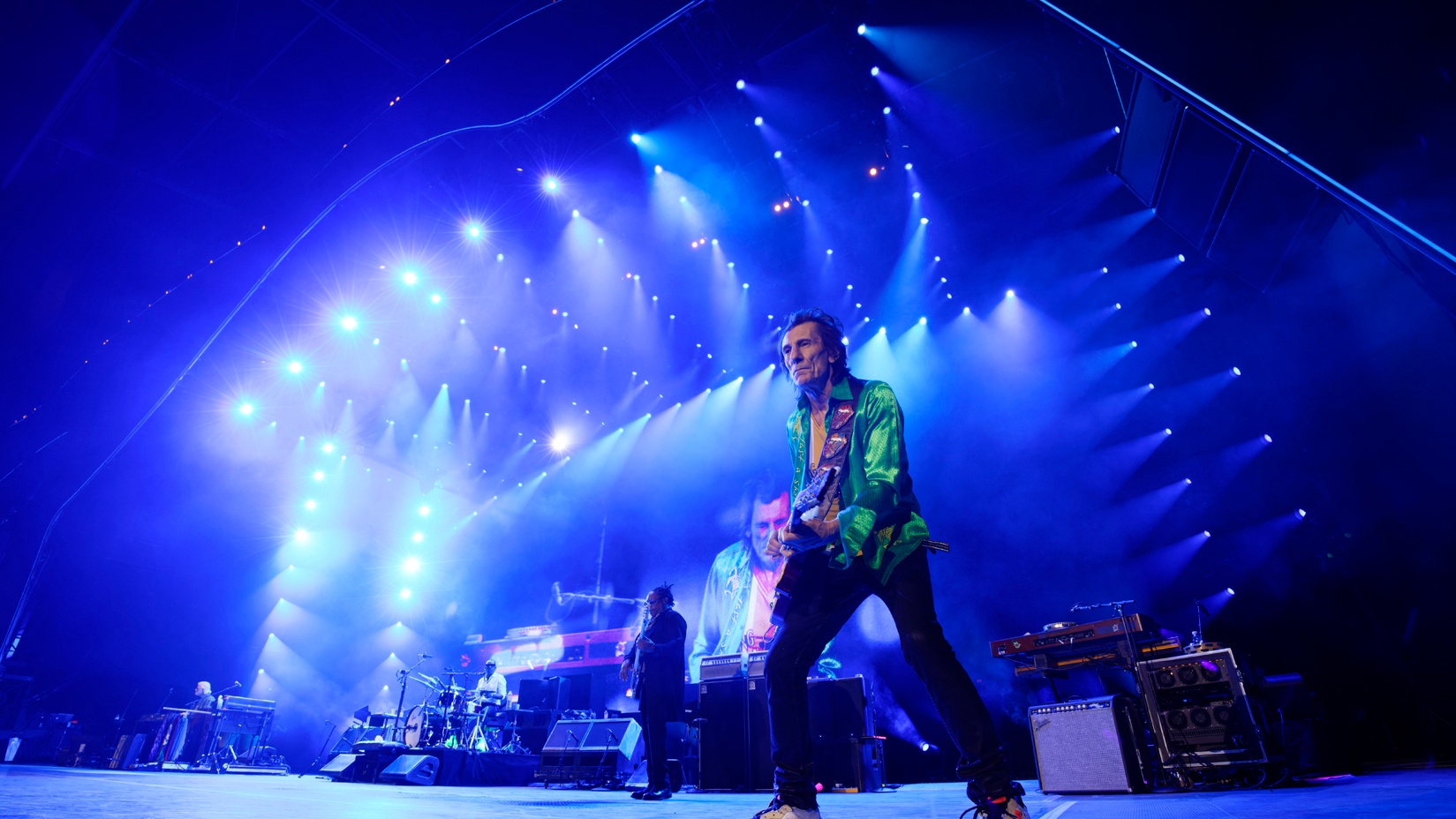 Ronnie Wood playing guitar on stage with The Rolling Stones in front of blue lighting and LED screen