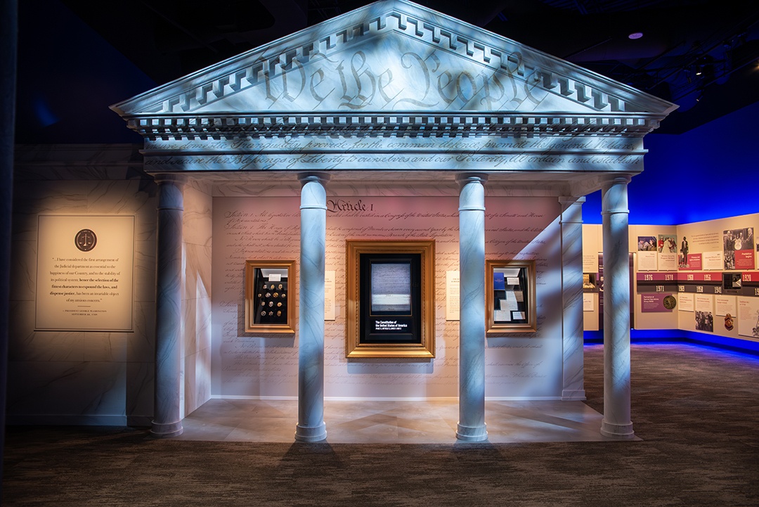 Museum display replica of white marbled, pillar buildings one might see in D.C. with We The People and other lettering from the US Constitution projected onto it with 3 interactive gold frames of history facts on the wall inside the building
