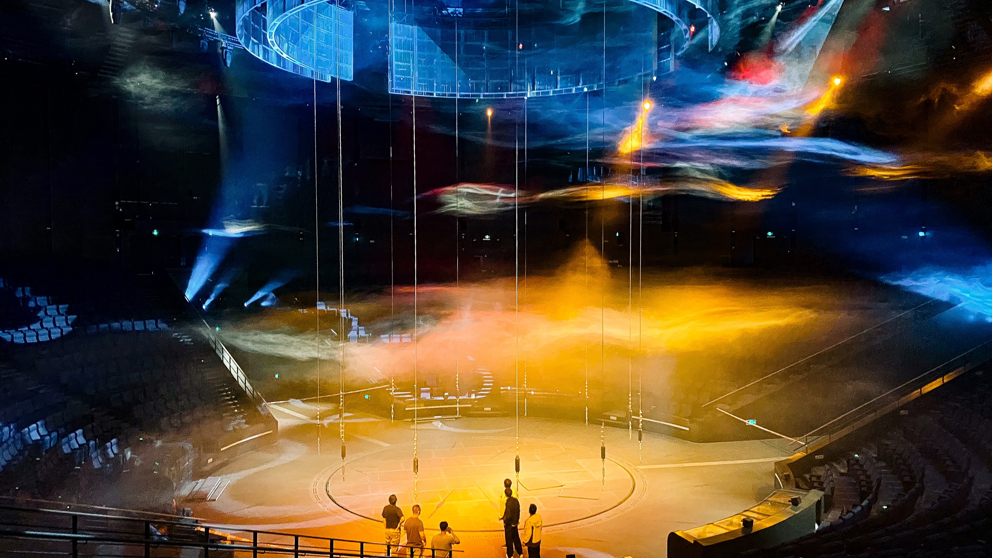 Multiple performer flying hoists hung down into the centre of a round theatre with lighting effects
