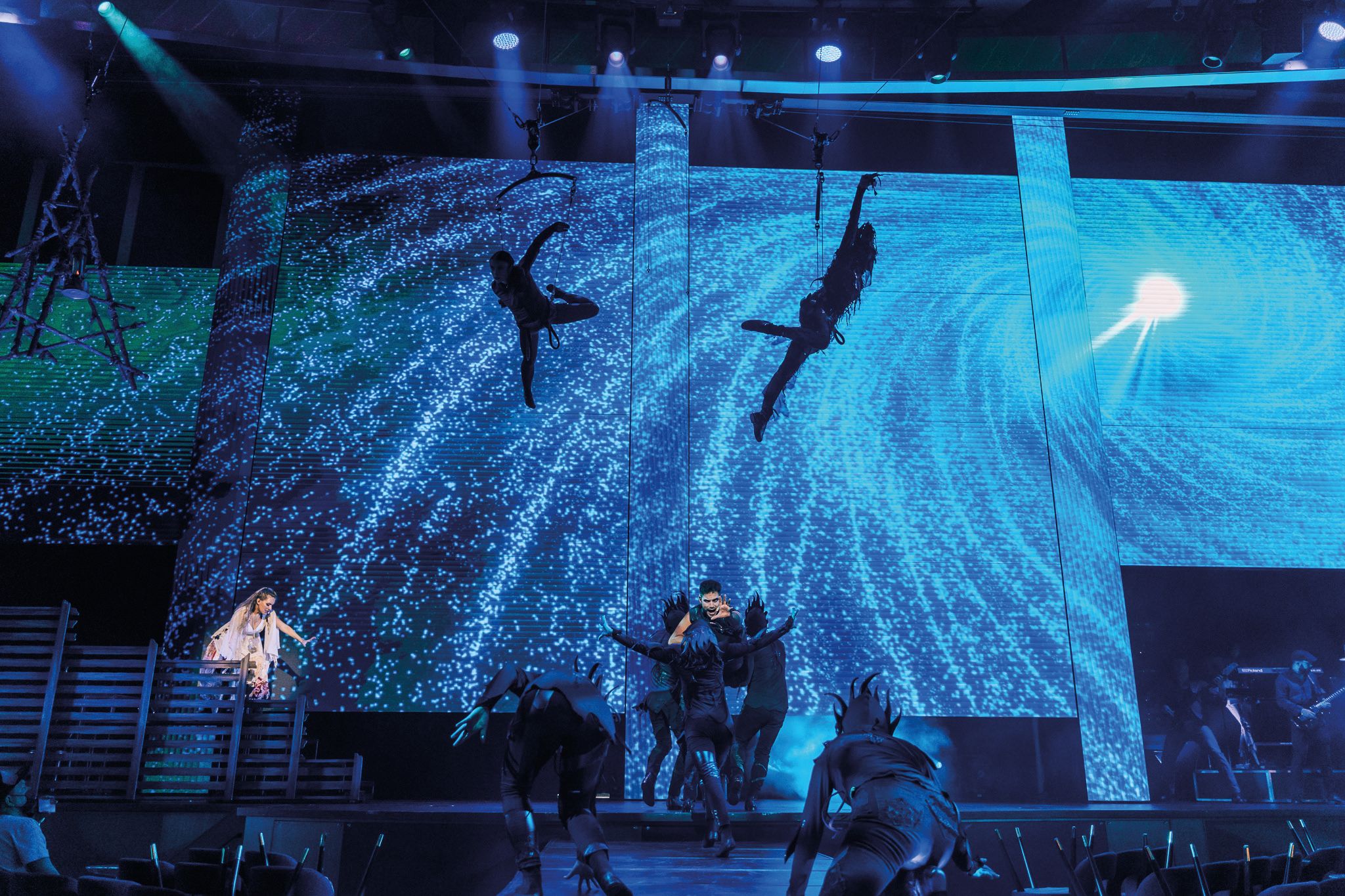 stage on a cruise ship with blue sparkles swirling on an LED screen that spans in entire stage with performers on stage and two performers flying high above