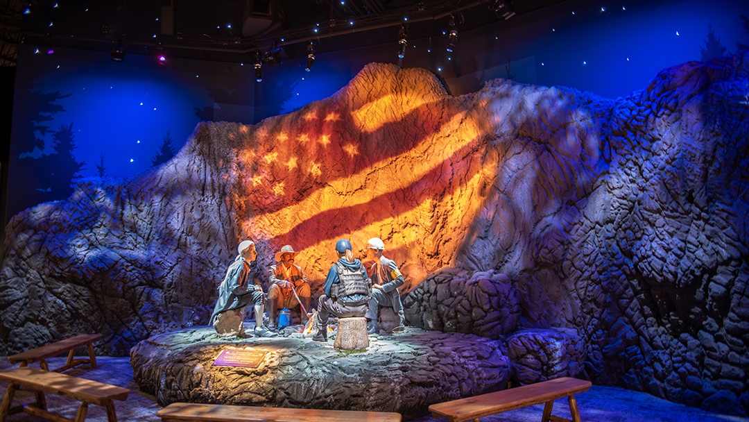 Museum experience with 4 realistic statue people sitting around a campfire with an American flag projected onto the large rockface behind them