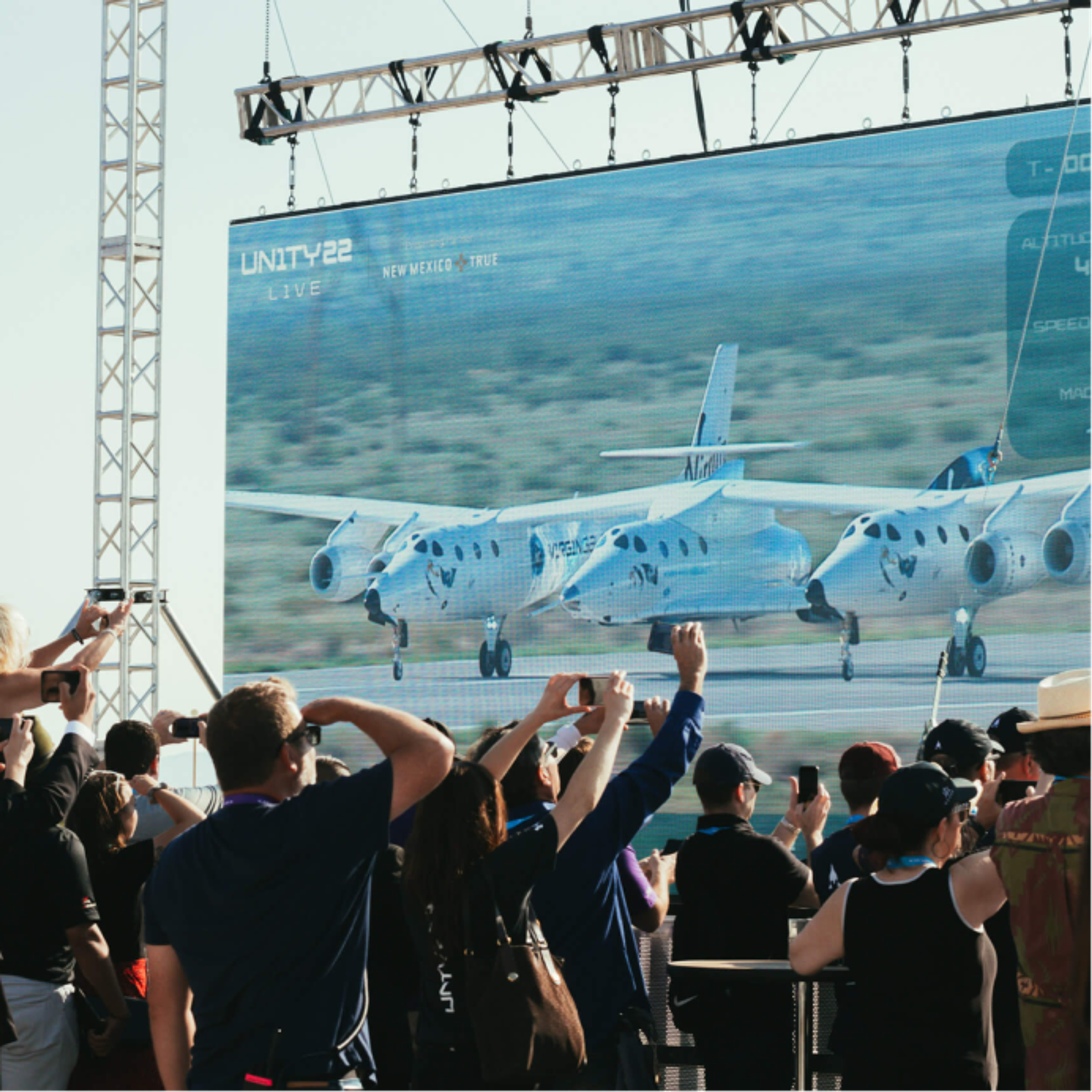 Virgin Galactic Launch is broadcast on a large LED screen, a crowd is in the foreground watching the broadcast