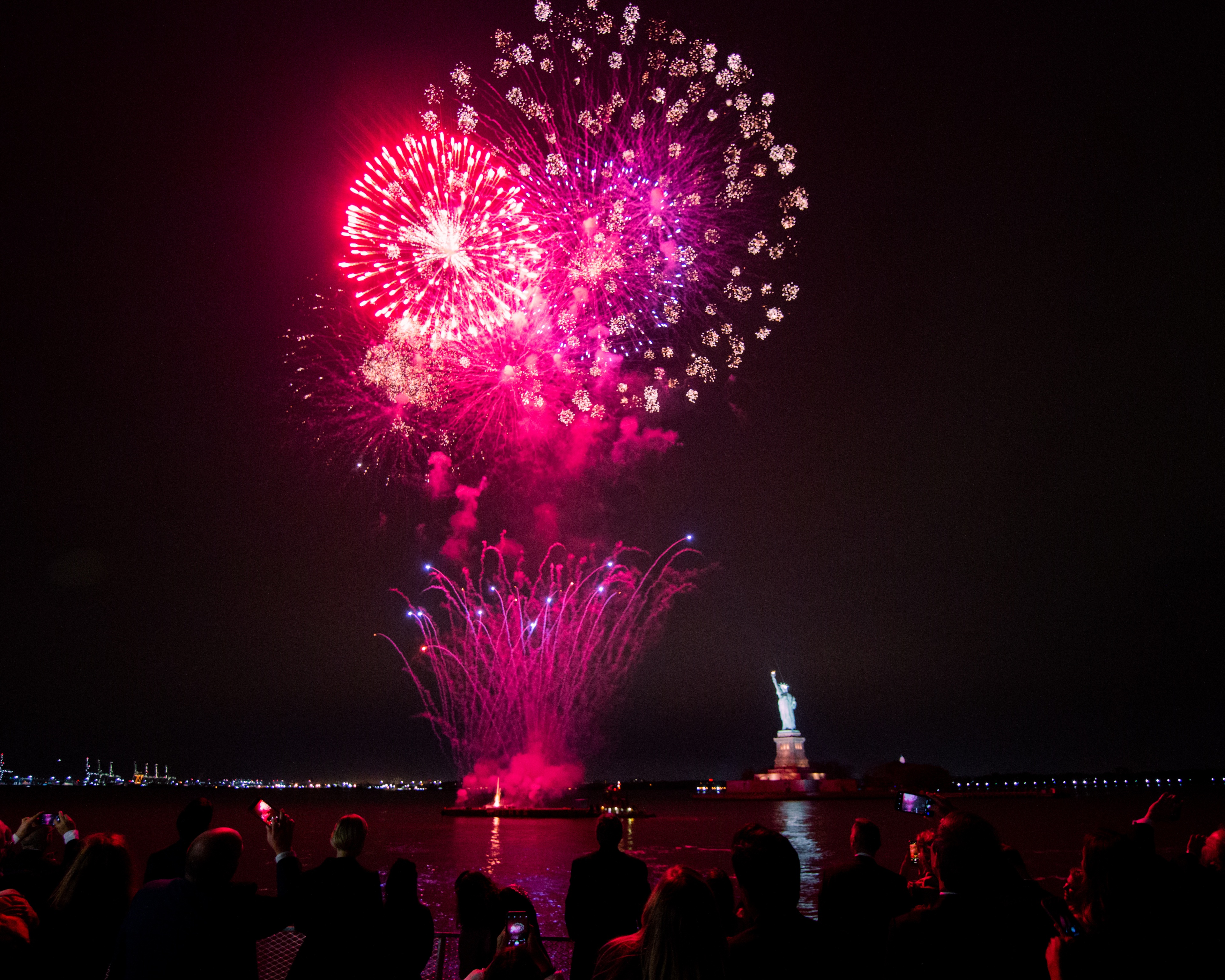 Huge firework display at night on river in front of the statue of liberty with crowd watching