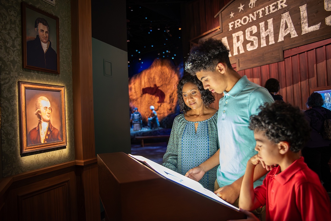 Museum with Frontier Marshalls in text in the background with 3 museum-goers captivated by an interactive digital display in the foreground and to the left there are a couple of framed US Presidents on the wall