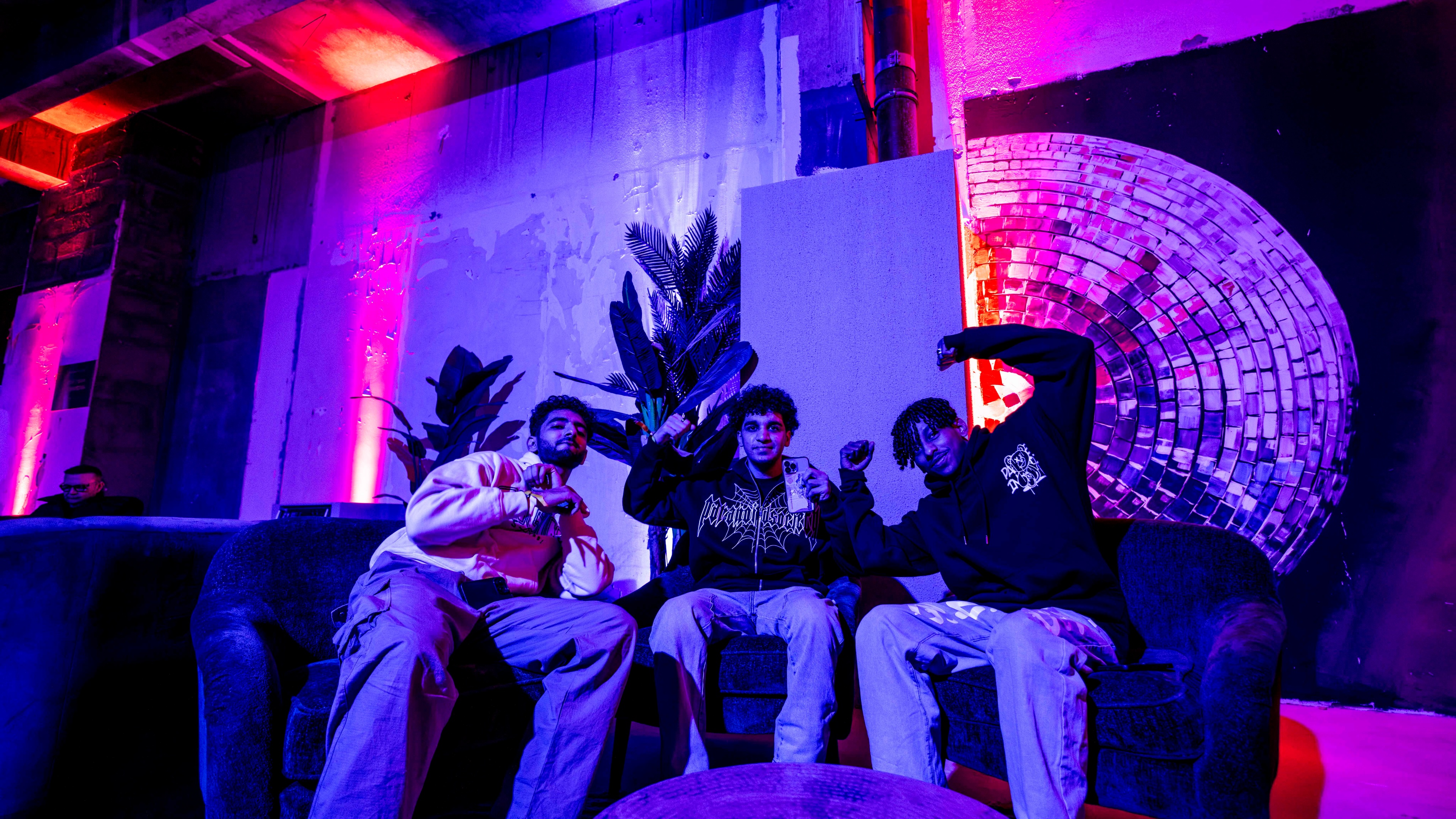 3 men sitting on couches signalling to the camera in dim lit industrial environment in front of disco ball and red lights