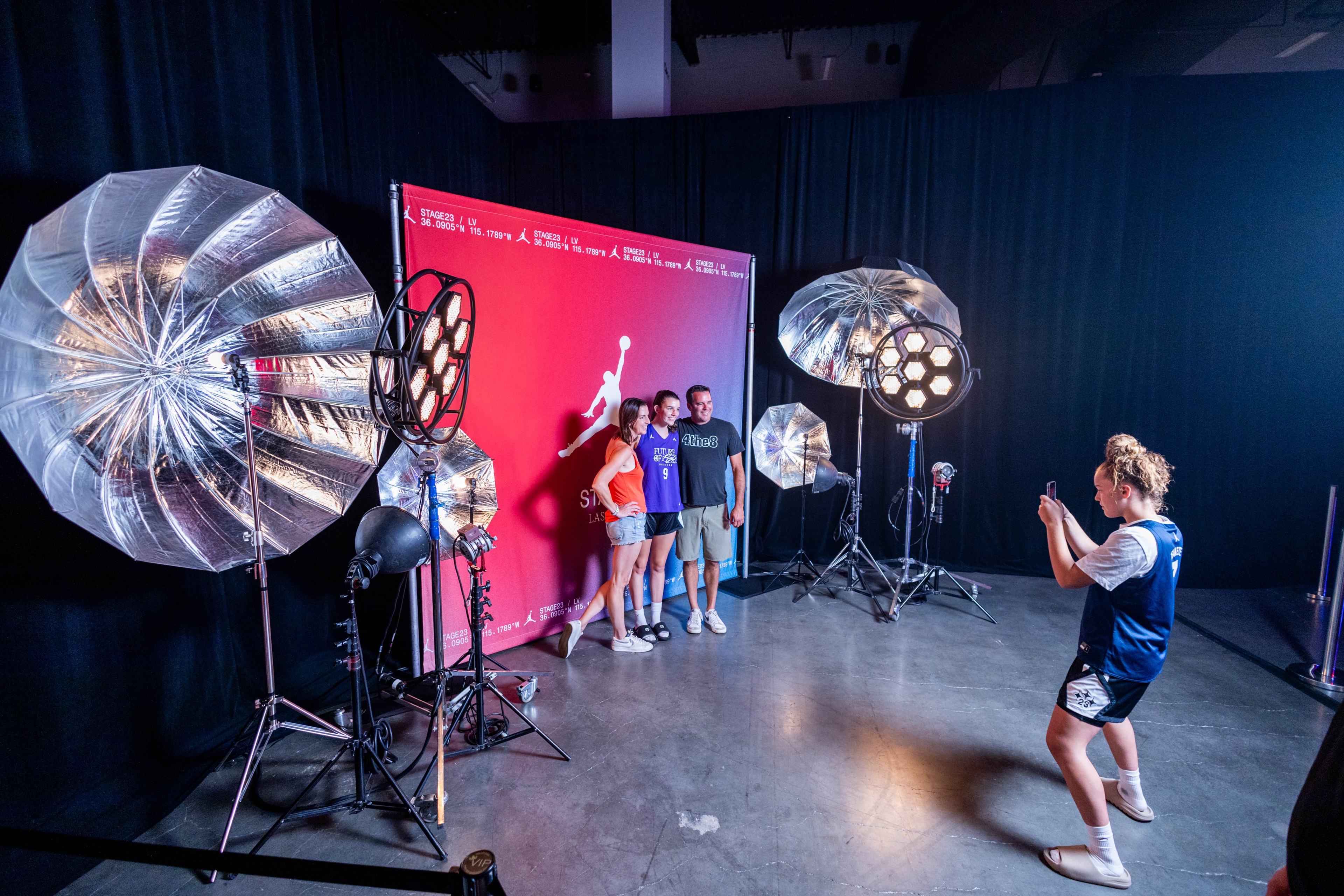 Nike Jordan Brand WNBA All-Star Weekend Las Vegas female in basketball attire taking photo of two females and one male standing in front of an event branded backdrop, with photography lamp equipment to the sides