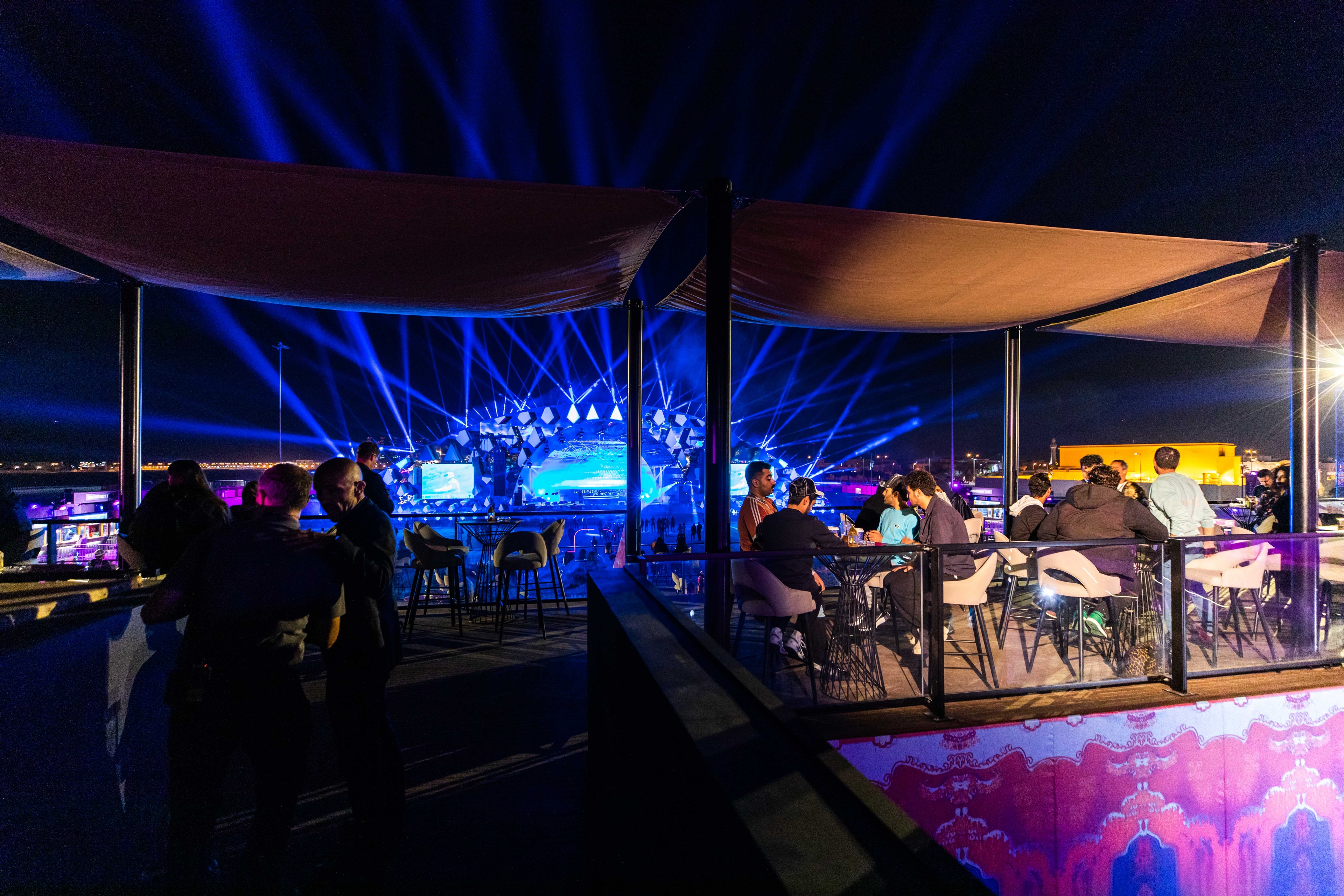Aravia MDLBEAST men sitting around rounded tables chatting and drinking in the balcony, concert happening in the backdrop