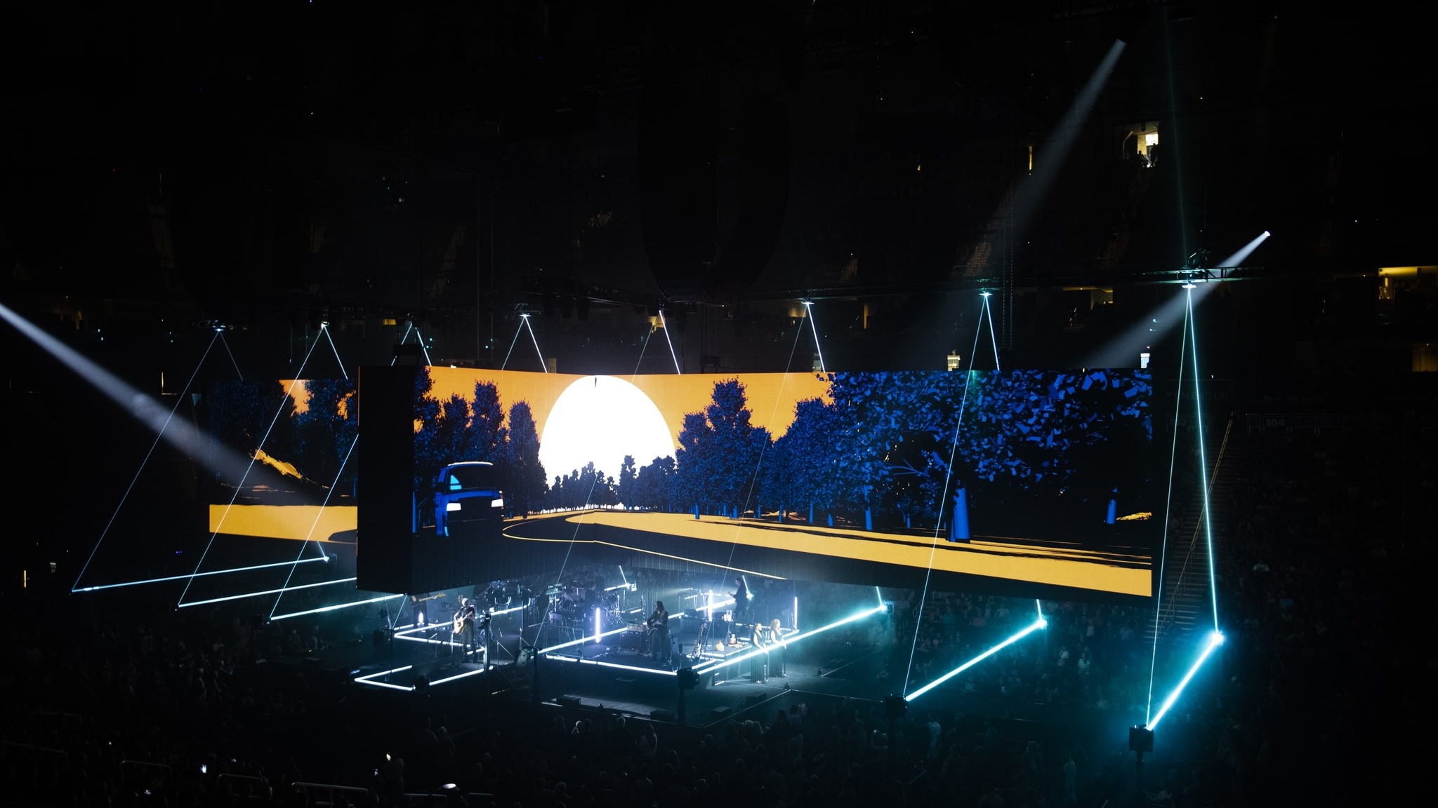 bird's eye view of Roger Waters This Is Not A Drill Tour stage featuring dimensional outlined triangles made of lights, screens show an illustration of a blue car on the road, surrounded by blue trees, a yellow sky, during sunset