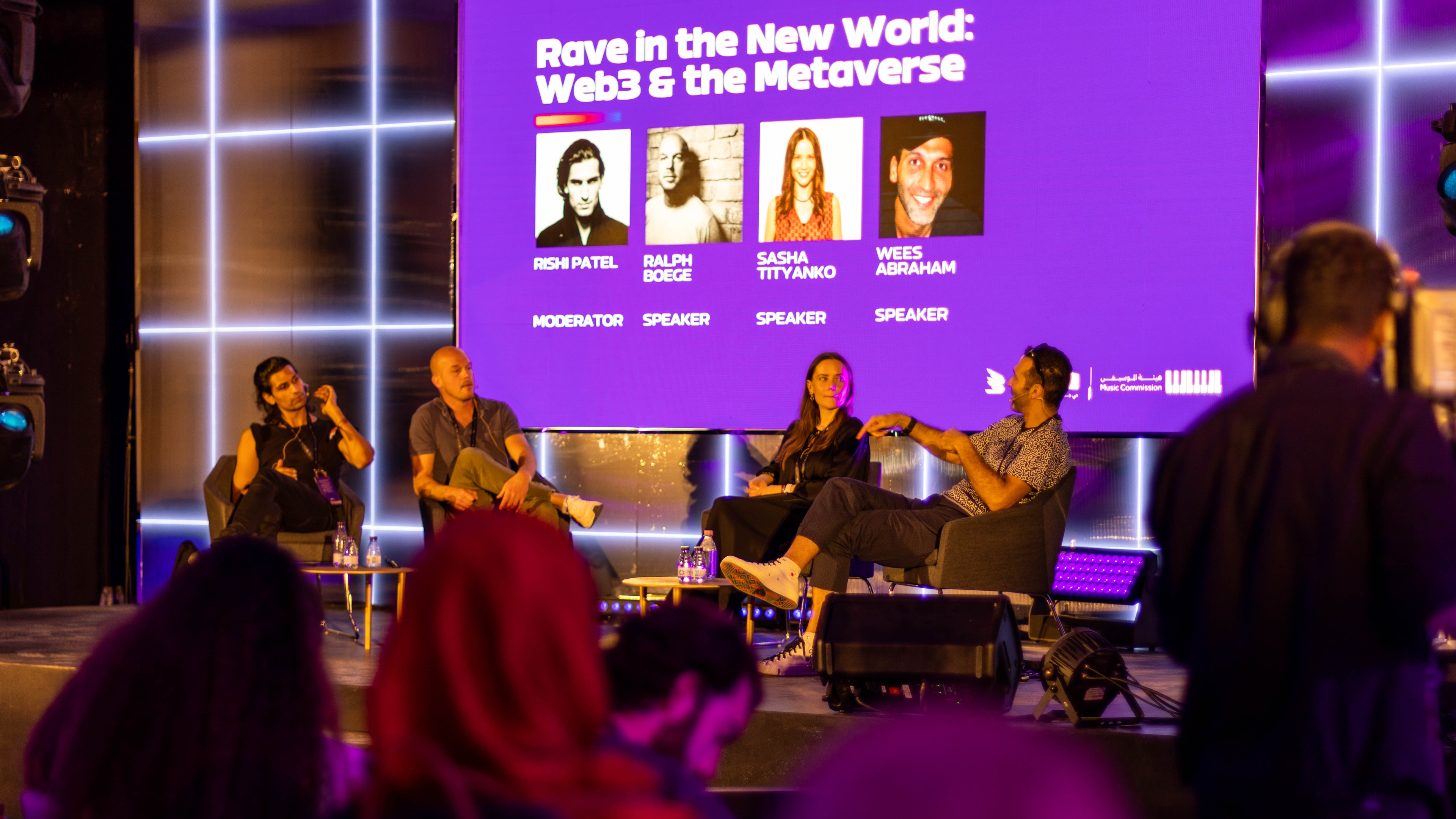 Four speakers sitting on chairs on stage, a screen behind them has the text "Rave in the New World: Web3 & The Metaverse"