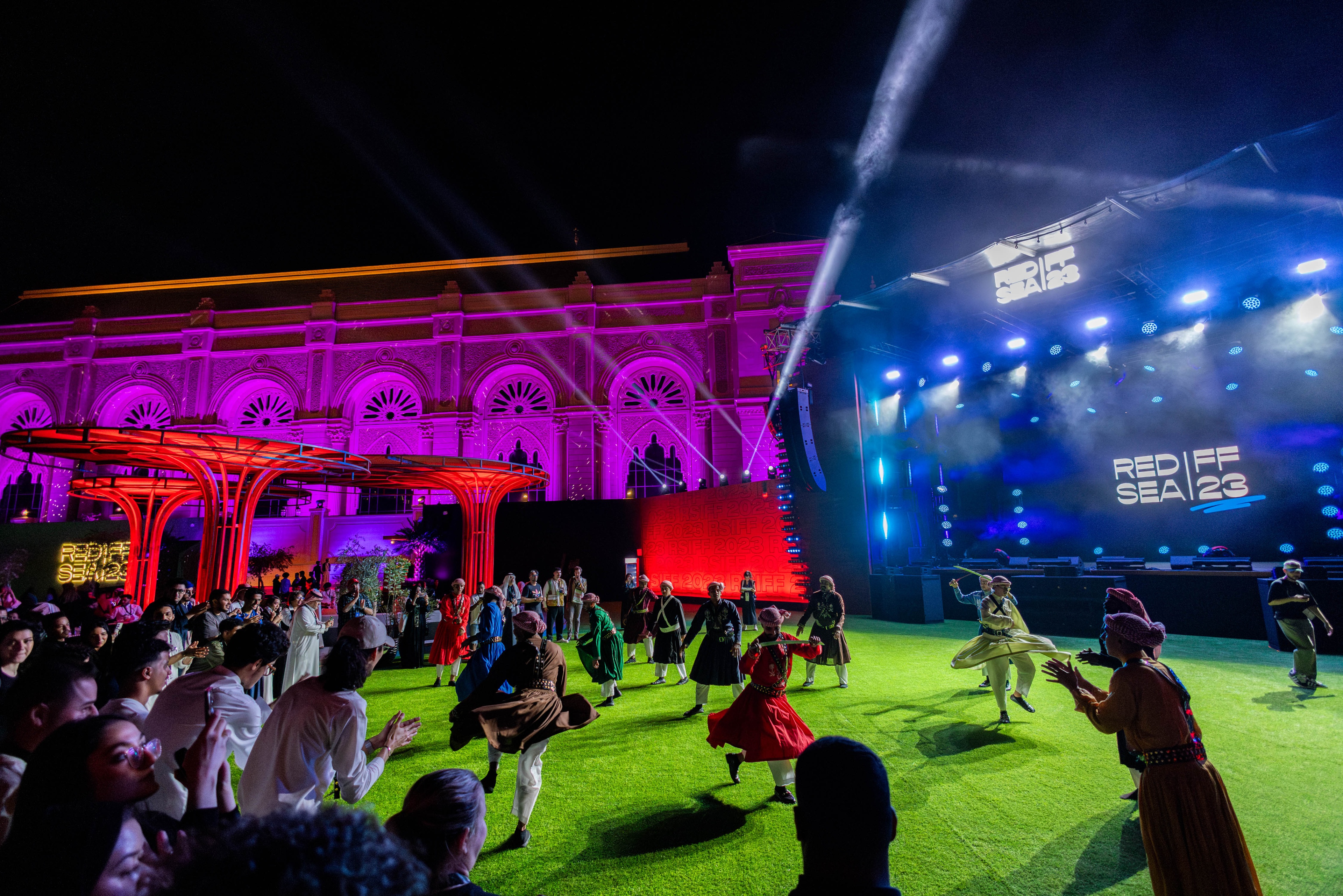 Dancers performing for crowd on green turf with pink and blue lights at night