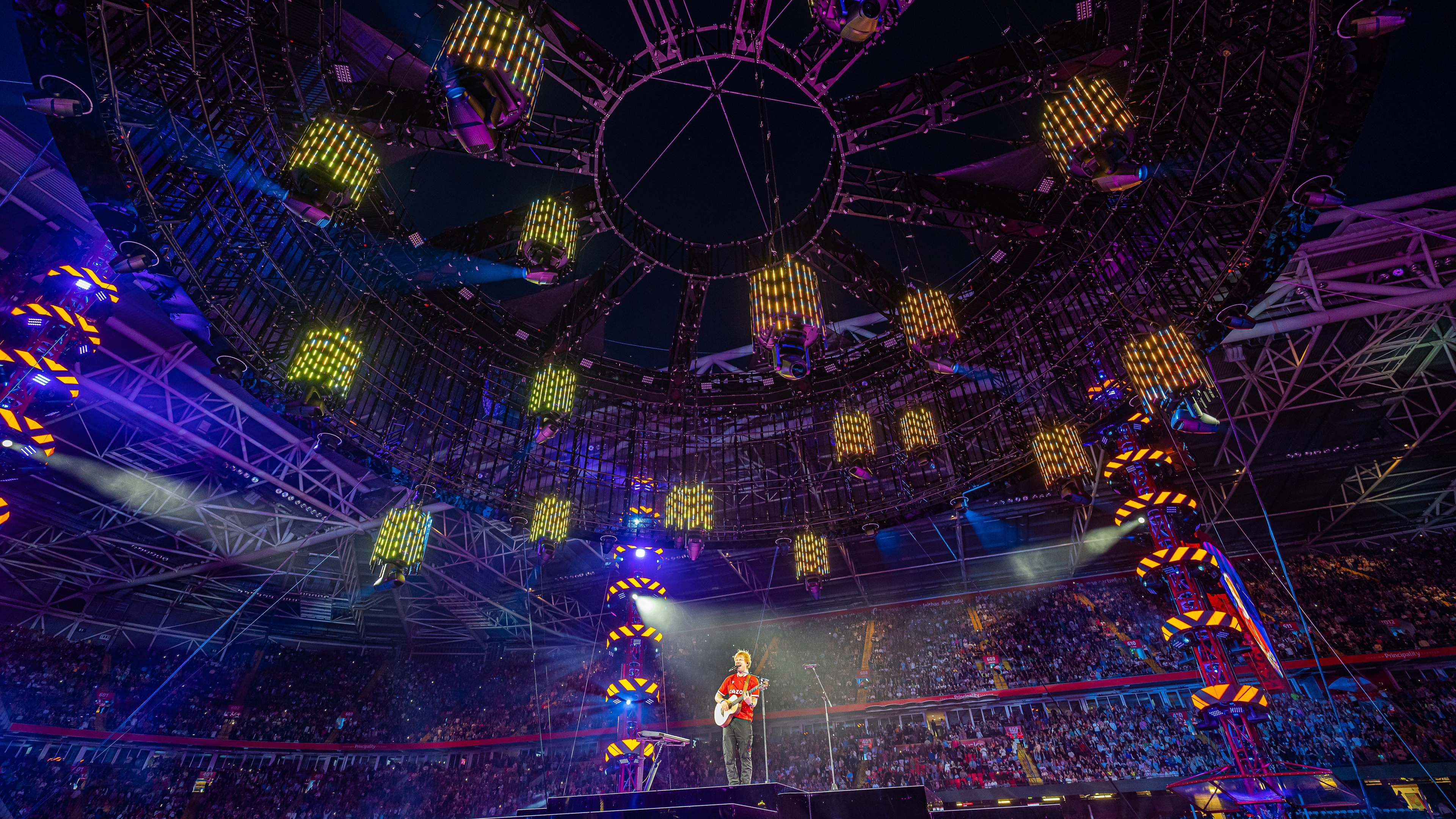 Ed Sheeran performing in red shirt on stage with purple and blue lights