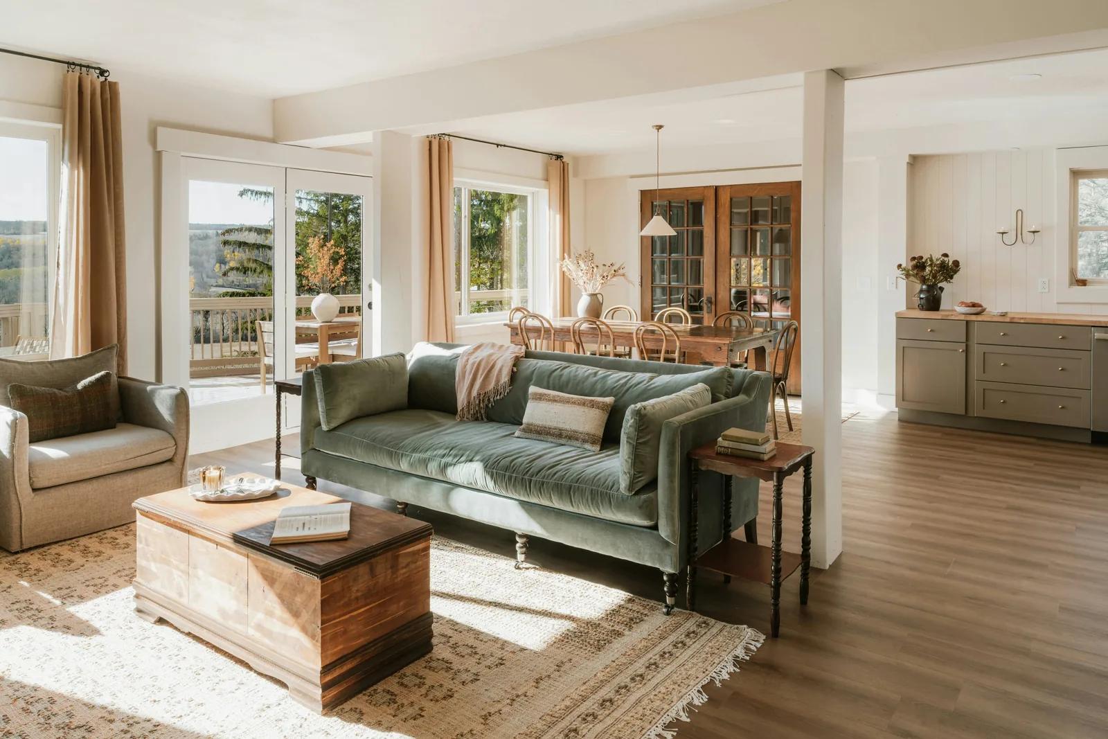 A sunlit open-plan living room with a sage velvet sofa, a wool throw folded over its arm, a solid wood chest used as a coffee table and a layered rug, with a dining room and warm timber doors beyond