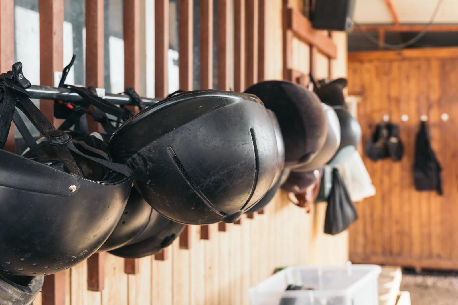 A row of well-worn black riding helmets hanging by their harness straps along a stable rail, with a saddle and tack on the timber wall behind