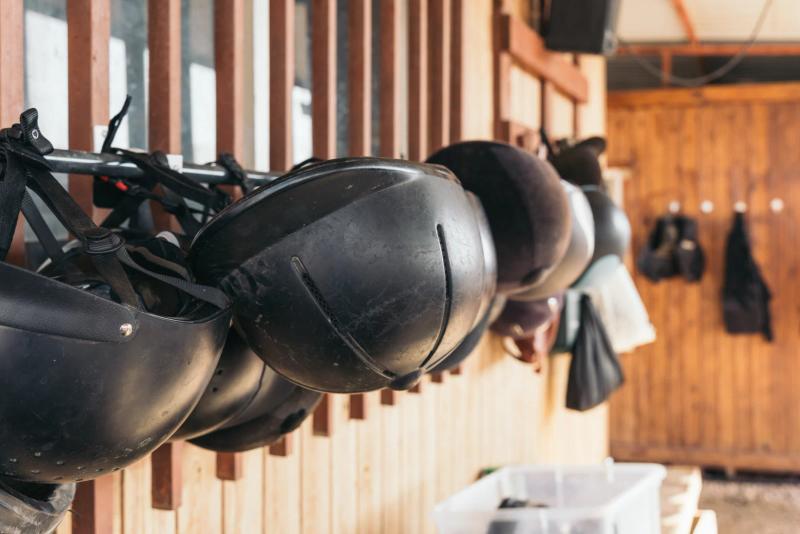 A row of well-worn black riding helmets hanging by their harness straps along a stable rail, with a saddle and tack on the timber wall behind