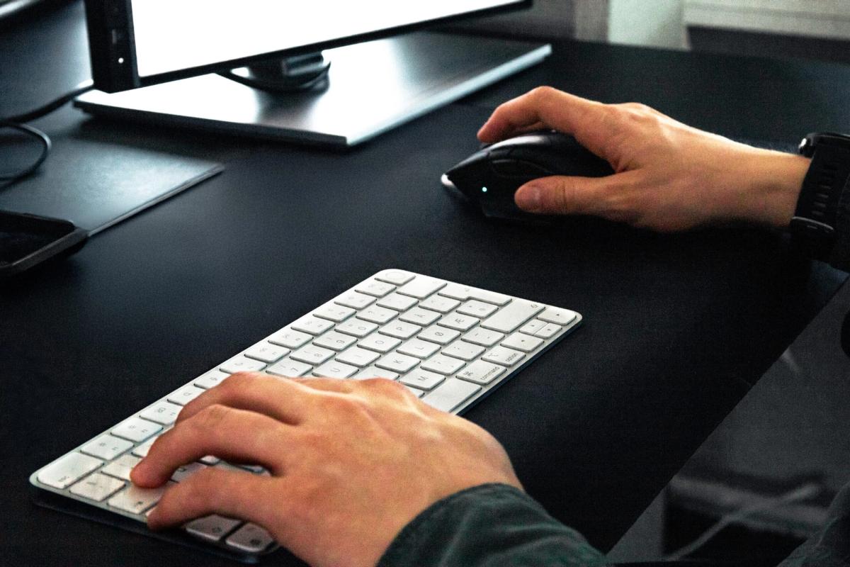 Nærbilde av hender på tastatur og mus ved et mørkt skrivebord