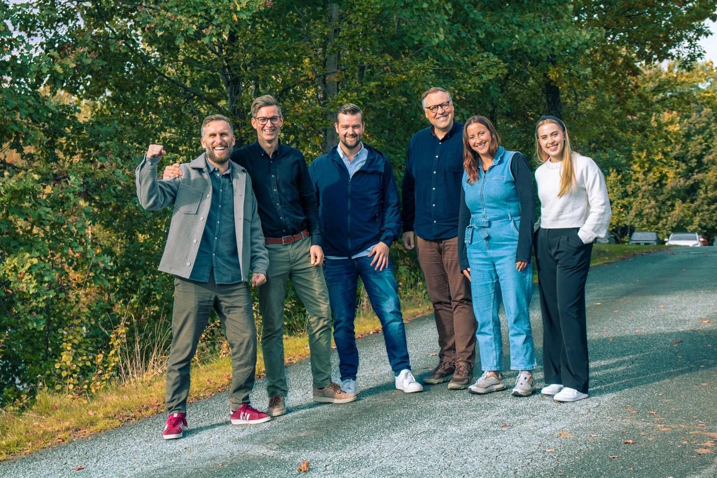 Seks kollegaer står samlet på en grusvei omgitt av høstfarget skog