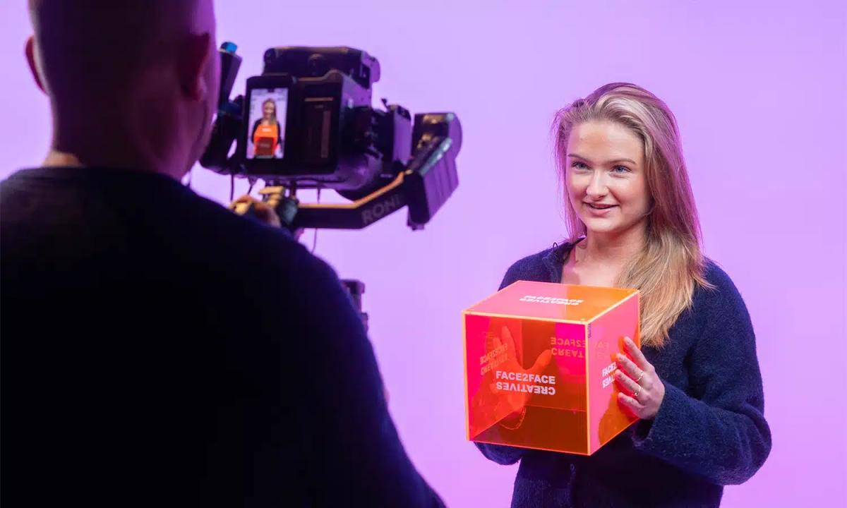 Kvinne holder en oransje eske foran et filmkamera i lilla studiolys