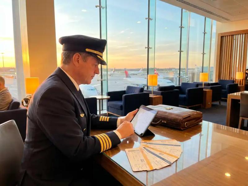 Airline captain reviewing flight itinerary on tablet in airport lounge, boarding passes fanned on surface, aircraft visible through floor-to-ceiling windows.