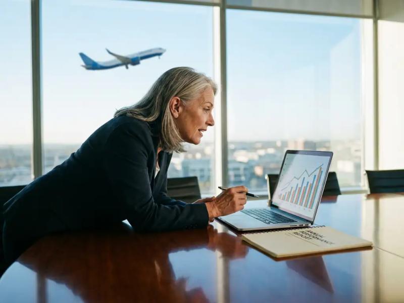 Senior female executive reviews upward-trending performance charts on laptop at conference table, aircraft visible through window behind her.
