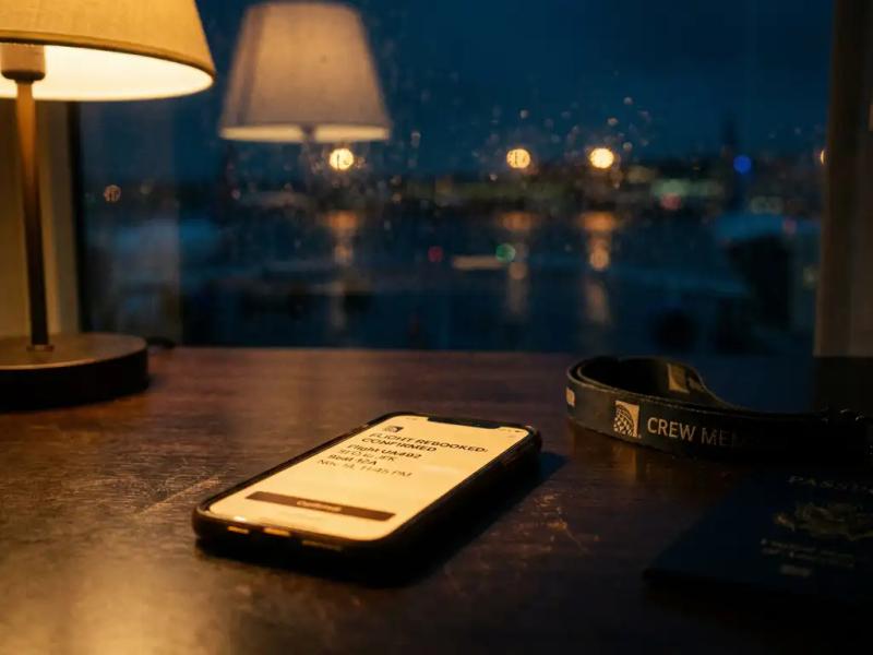 Glowing smartphone showing flight rebooking confirmation on dark wooden desk beside crew lanyard and passport, rain-streaked airport window at midnight.