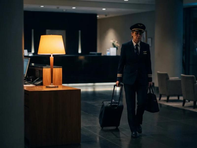 Tired airline crew member in uniform rolling a suitcase through a dimly lit hotel lobby at night, warm amber lamp glowing at reception desk.