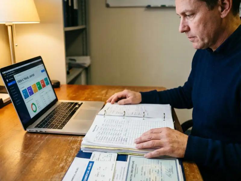 Maritime operations manager reviewing crew scheduling binder beside laptop displaying unified crew and travel dashboard on wooden desk