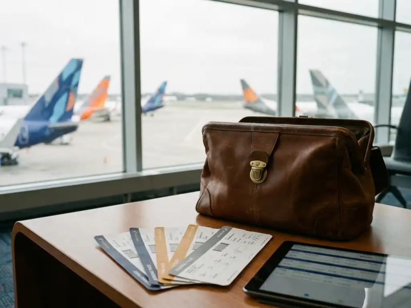 Worn leather pilot flight bag open on airport lounge table with boarding passes and crew scheduling tablet, parked aircraft visible through terminal windows.