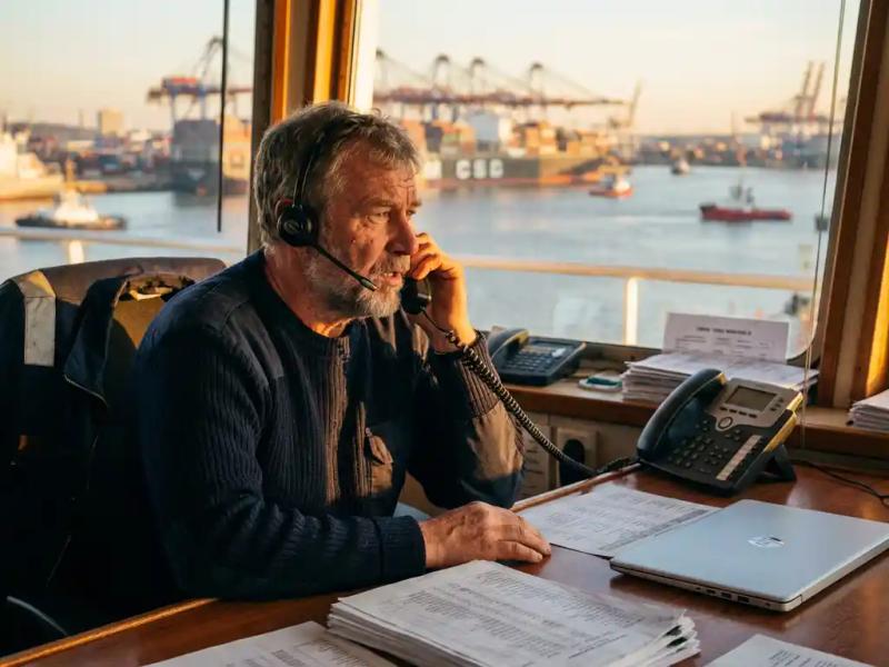 Maritime crew manager at port authority desk reviewing vessel schedules via headset, container ships visible through harbor window behind him.