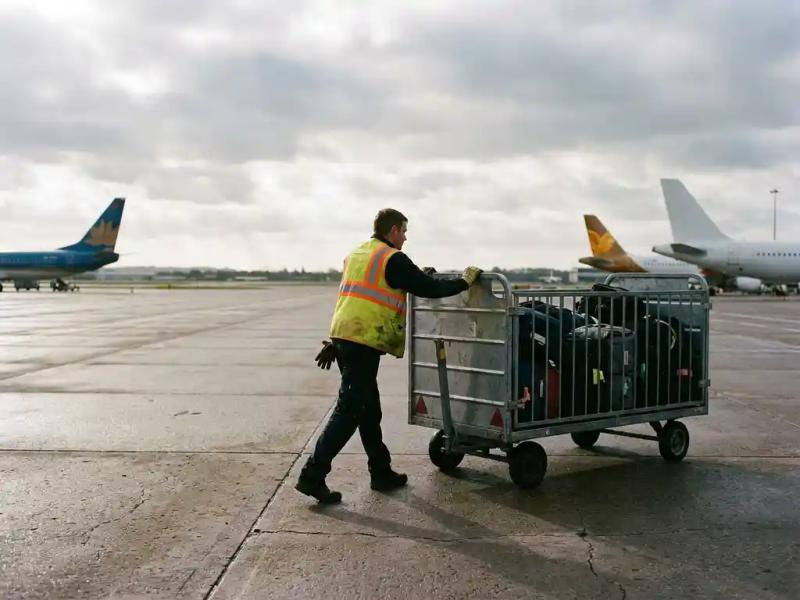 Ground handler in high-visibility vest rolling a luggage trolley across airport tarmac, aircraft tails visible in the background.