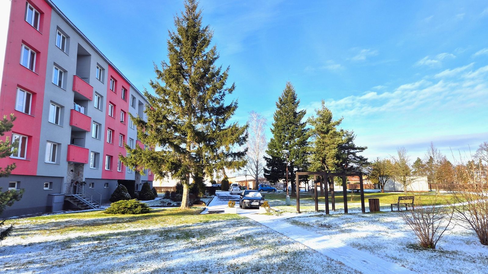 Bytový dům se šedou a červenou fasádou u zasněženého parku se vzrostlými smrky