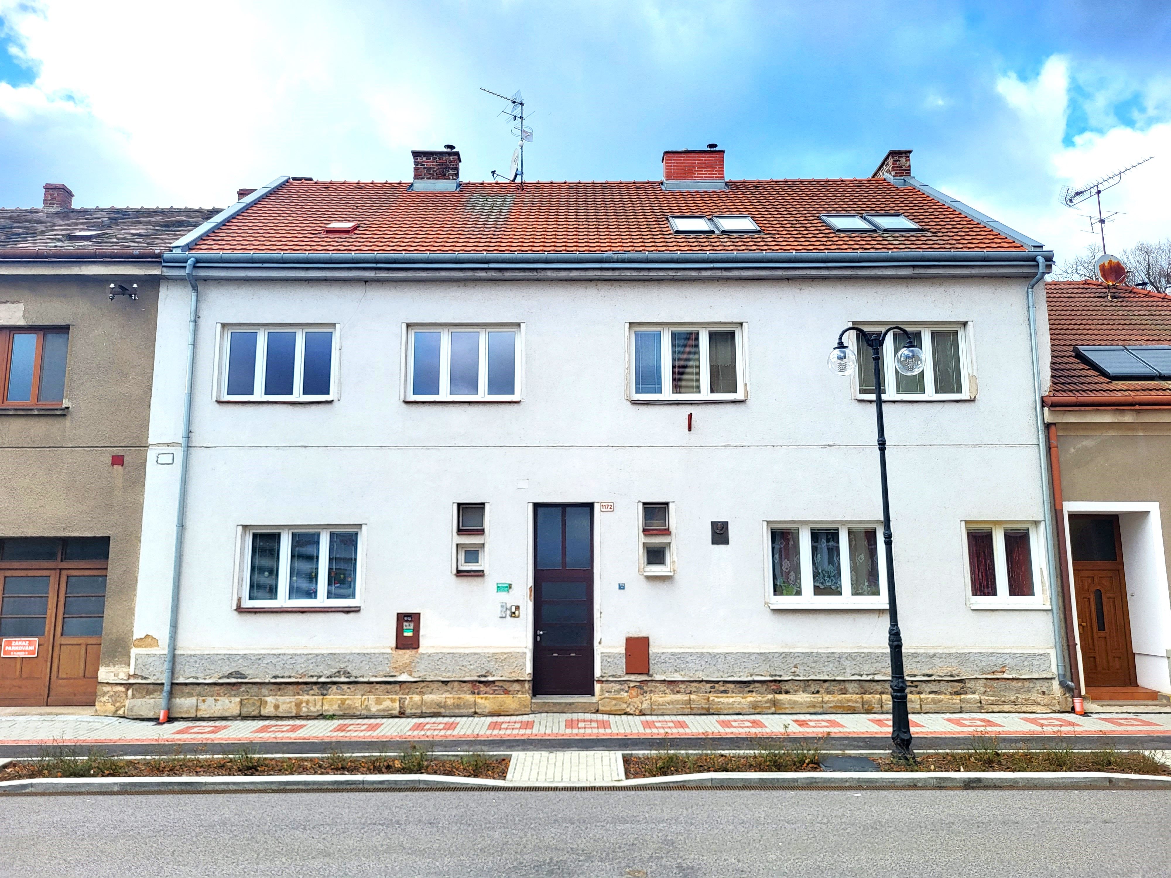 Bílý patrový řadový dům s červenou taškovou střechou u ulice s pouliční lampou