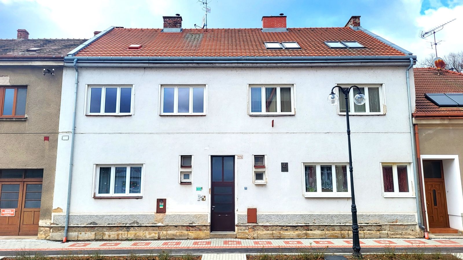 Bílý patrový řadový dům s červenou taškovou střechou u ulice s pouliční lampou