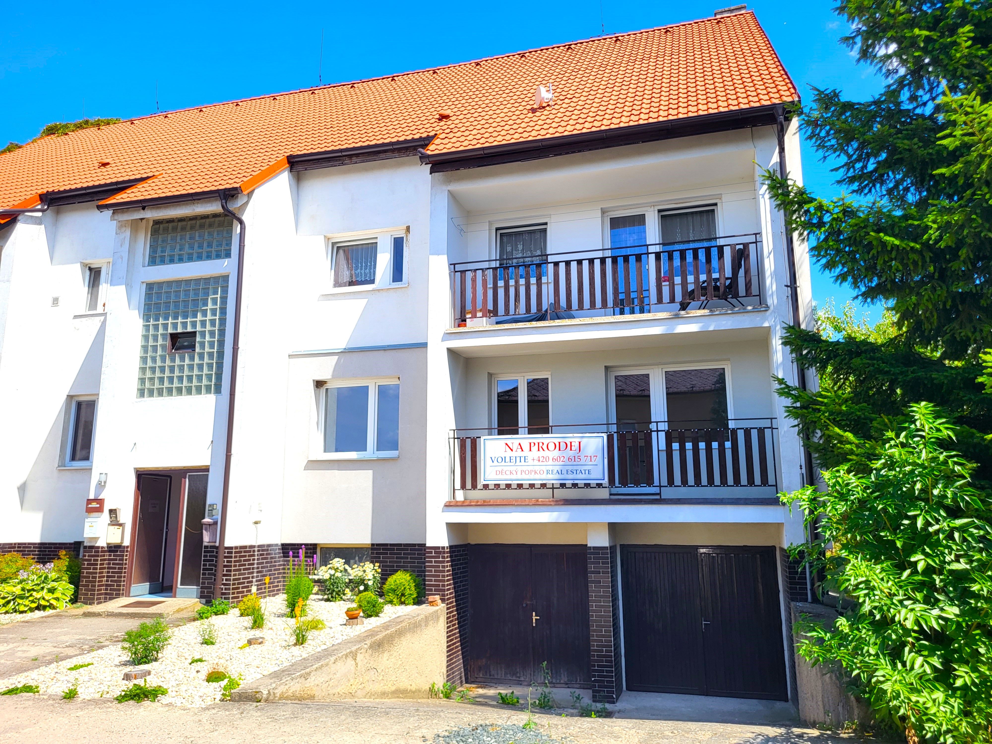 Bílý bytový dům s oranžovou střechou, balkony a garážemi v přízemí obklopený zelení