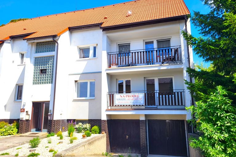 Bílý bytový dům s oranžovou střechou, balkony a garážemi v přízemí obklopený zelení