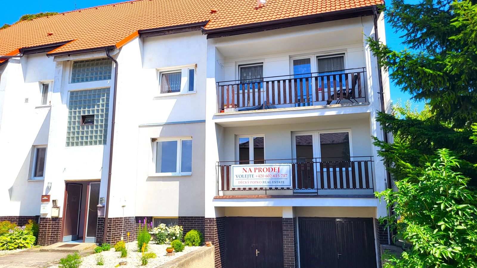 Bílý bytový dům s oranžovou střechou, balkony a garážemi v přízemí obklopený zelení