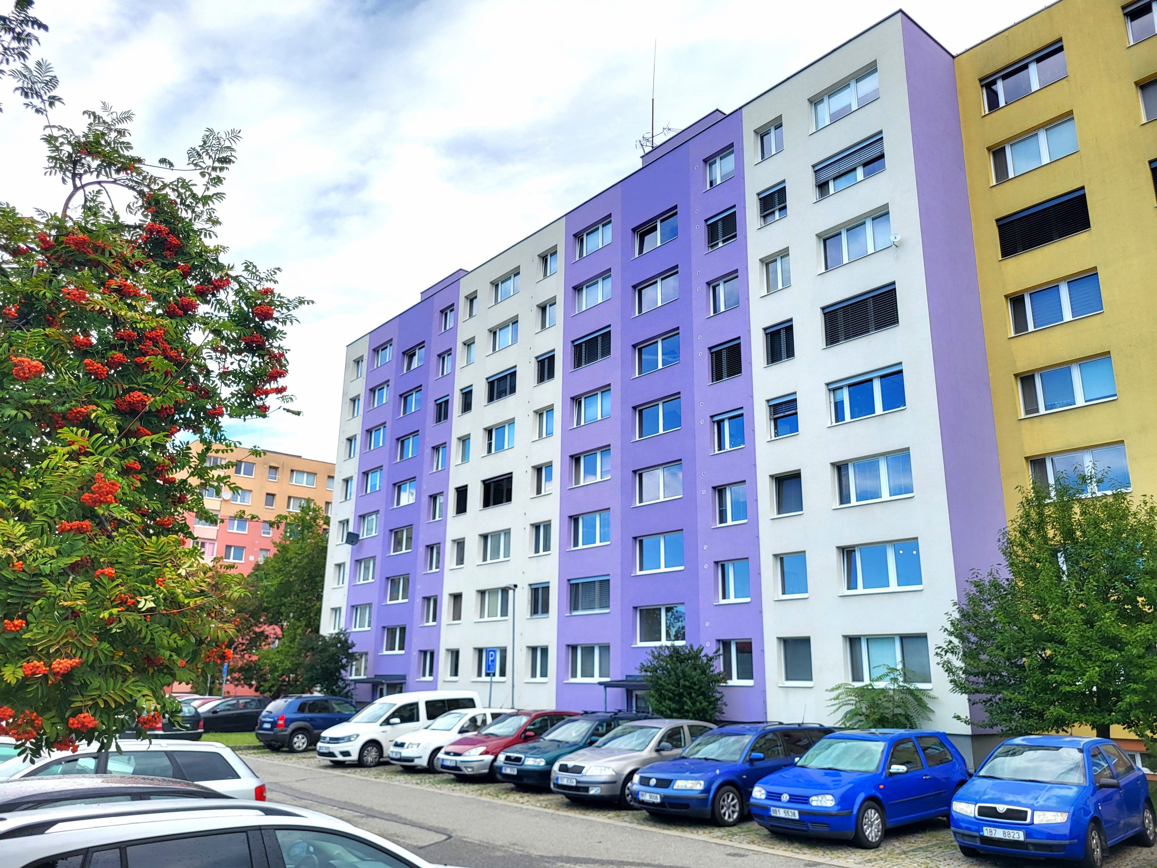 Panelový bytový dům s fialovou a bílou fasádou a parkovištěm s auty