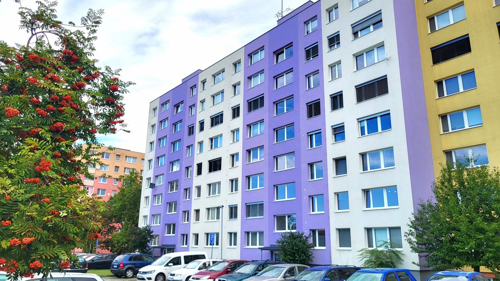 Panelový bytový dům s fialovou a bílou fasádou a parkovištěm s auty