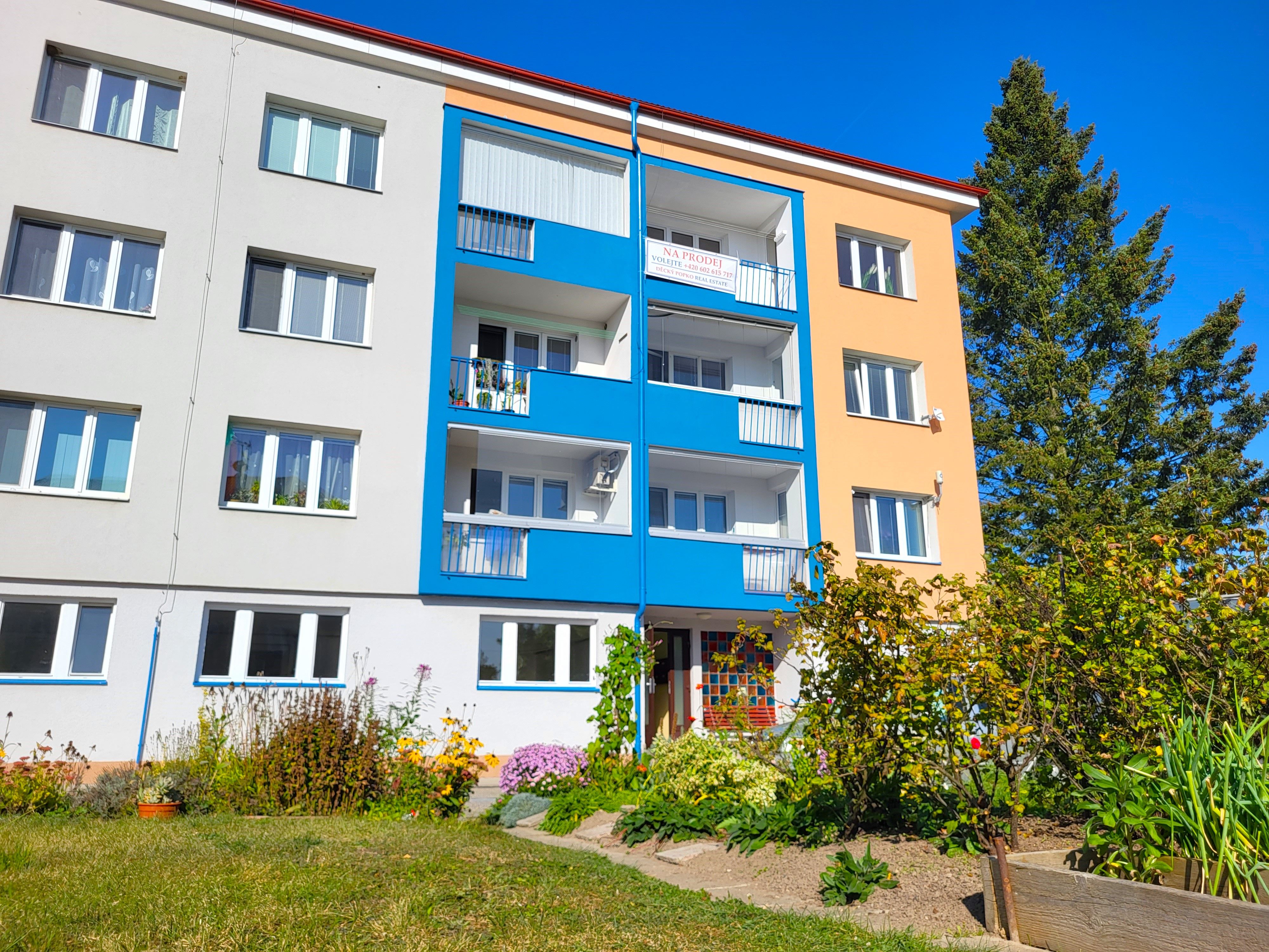 Bytový dům s modrými balkony a oranžovou fasádou a předzahrádkou plnou květin