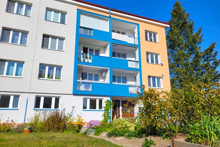 Bytový dům s modrými balkony a oranžovou fasádou a předzahrádkou plnou květin