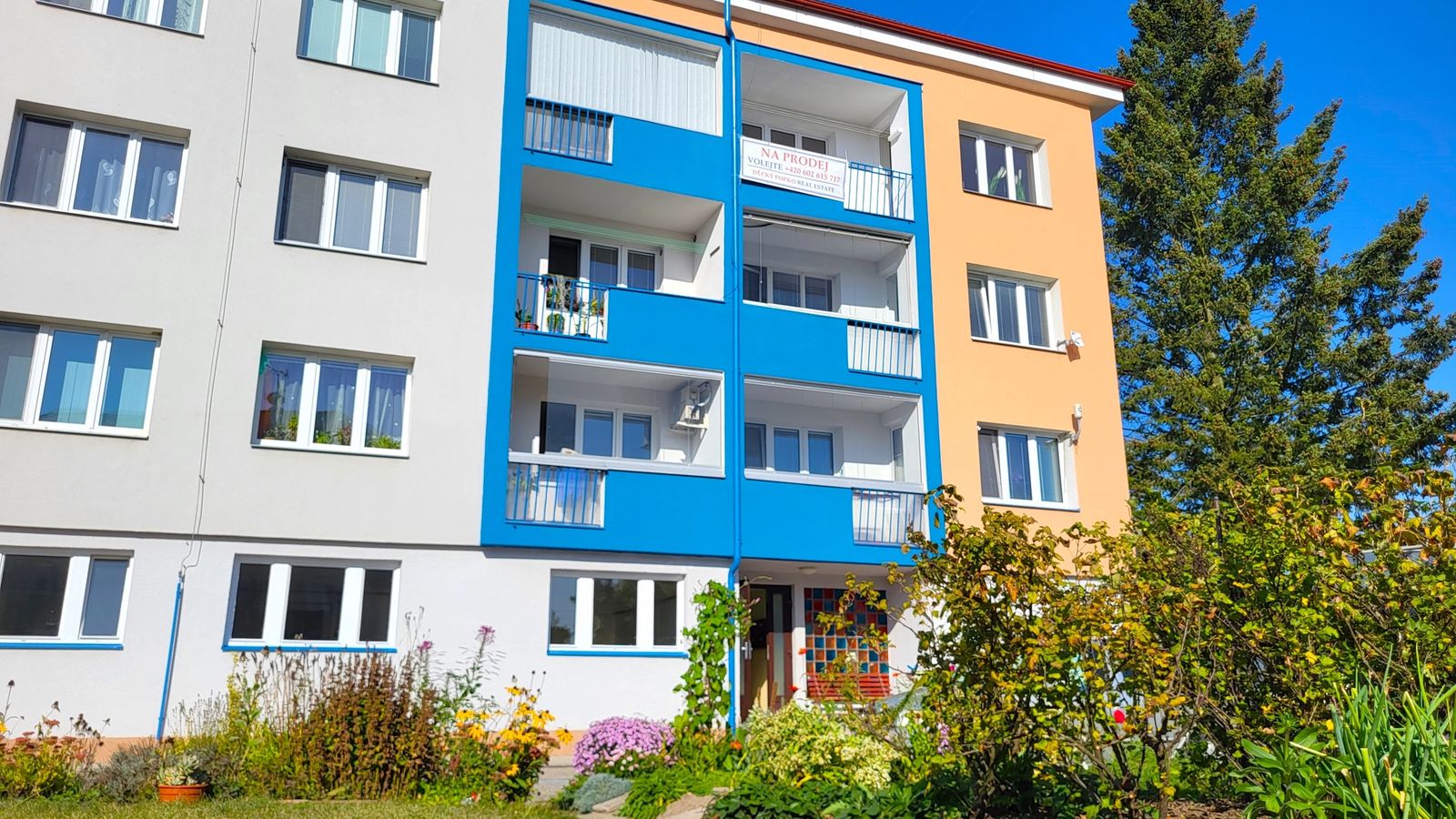Bytový dům s modrými balkony a oranžovou fasádou a předzahrádkou plnou květin