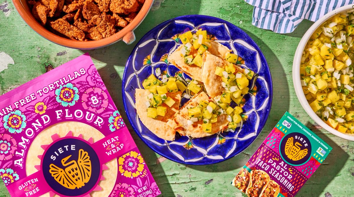 Siete Tortillas laying flat alongside Siete Al Pastor Taco Seasoning Packet. Bowls of Pork Al Pastor and Pineapple Pico de Gallo lay in the photo as well with a plate of Quesadillas Al Pastor at the center. Topped with Pineapple Pico de Gallo.