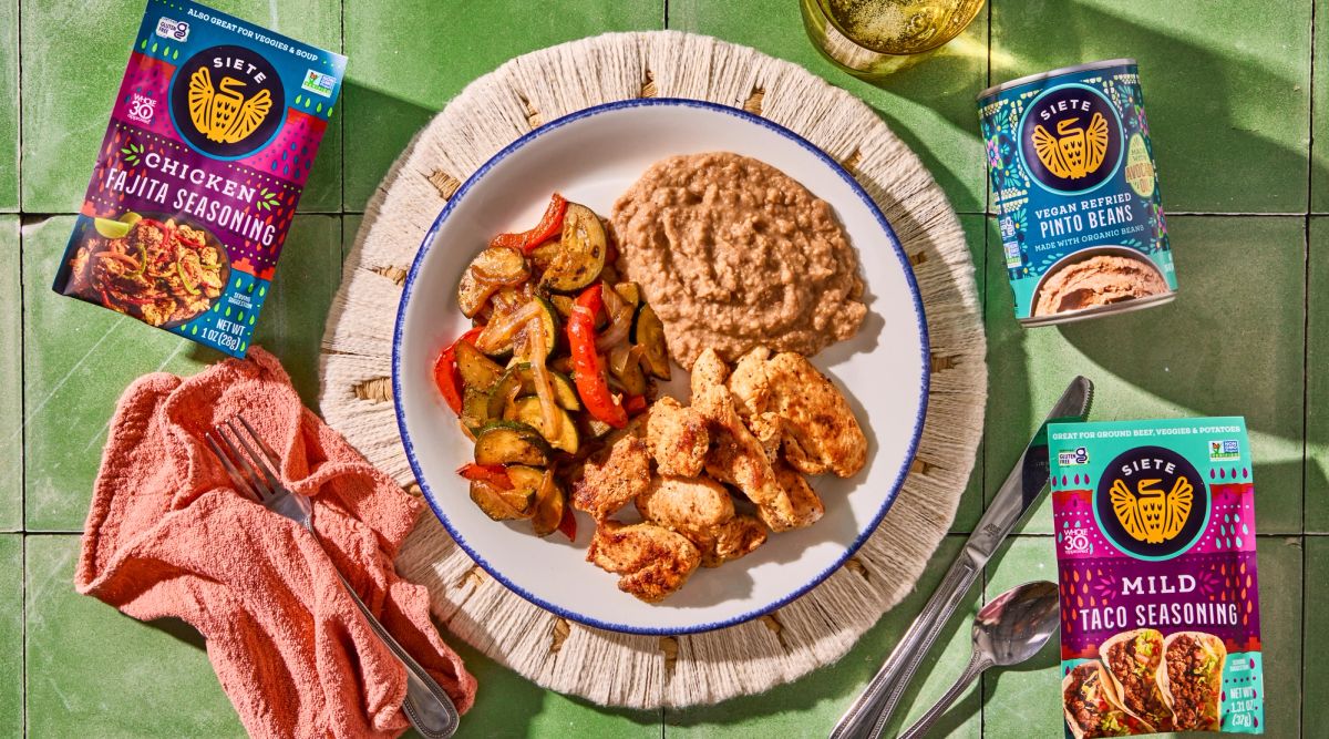 A plate of refried pinto beans, chicken fajita bites, and taco-seasoned veggies with Siete's Mild Taco Seasoning laying flat alongside Siete's Refried Pinto Beans and Siete's Chicken Fajita Seasoning