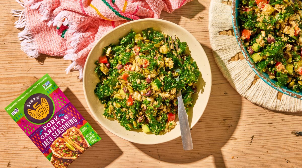 Kale and Quinoa Salad in a bowl with a fork in it. Set next to a packet of Siete Carnitas Seasoning laying flat. Pink towel underneath the bowl. 