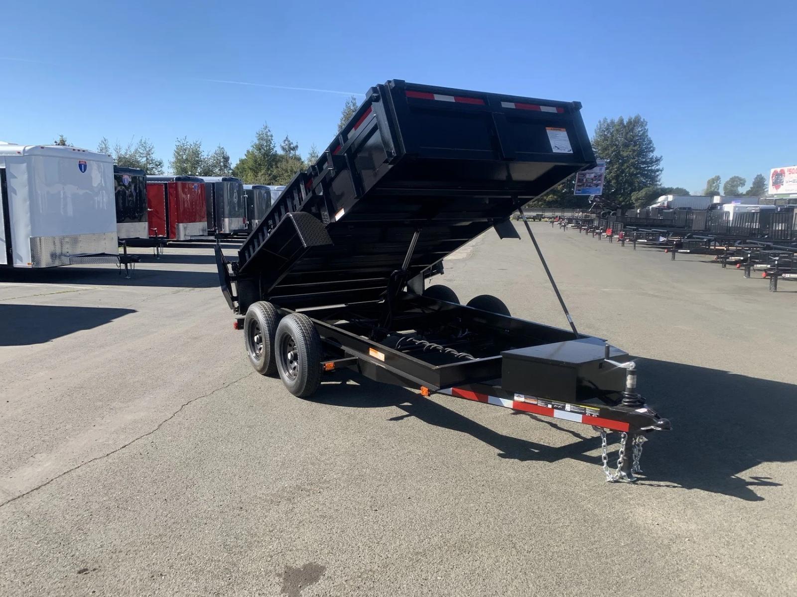 Black tandem-axle dump trailer with the bed tilted up at full dump angle, parked beside a row of enclosed cargo trailers on a sunny dealer lot